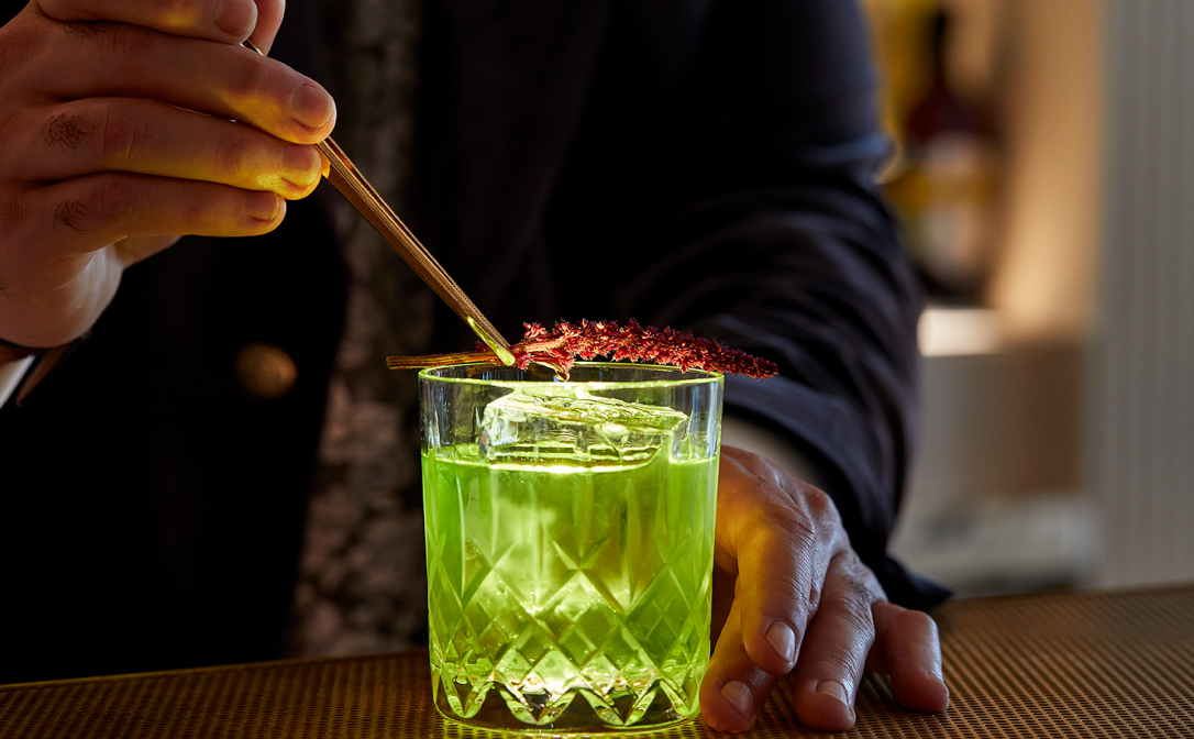 A bright green cocktail in a clear glass, garnished with a red flower
