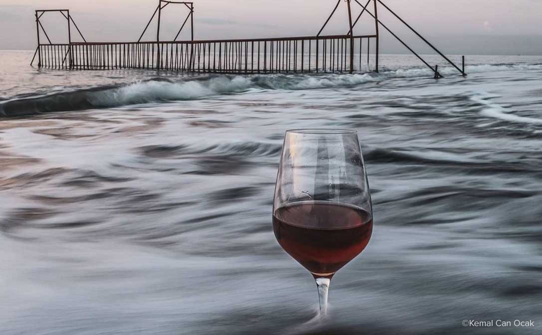 Photography by Kemal Can Ocak. A glass of red wine floating in the waves at sunrise